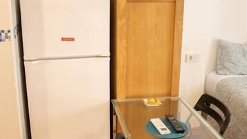 Una cocina compacta con un refrigerador blanco, un mueble de madera, una mesa de vidrio con sillas y un televisor fijado a la pared.