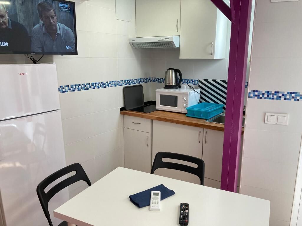 Een compacte keuken met een magnetron, waterkoker en een televisie boven het aanrecht. Er staat een tafel met stoelen en een kleine koelkast.