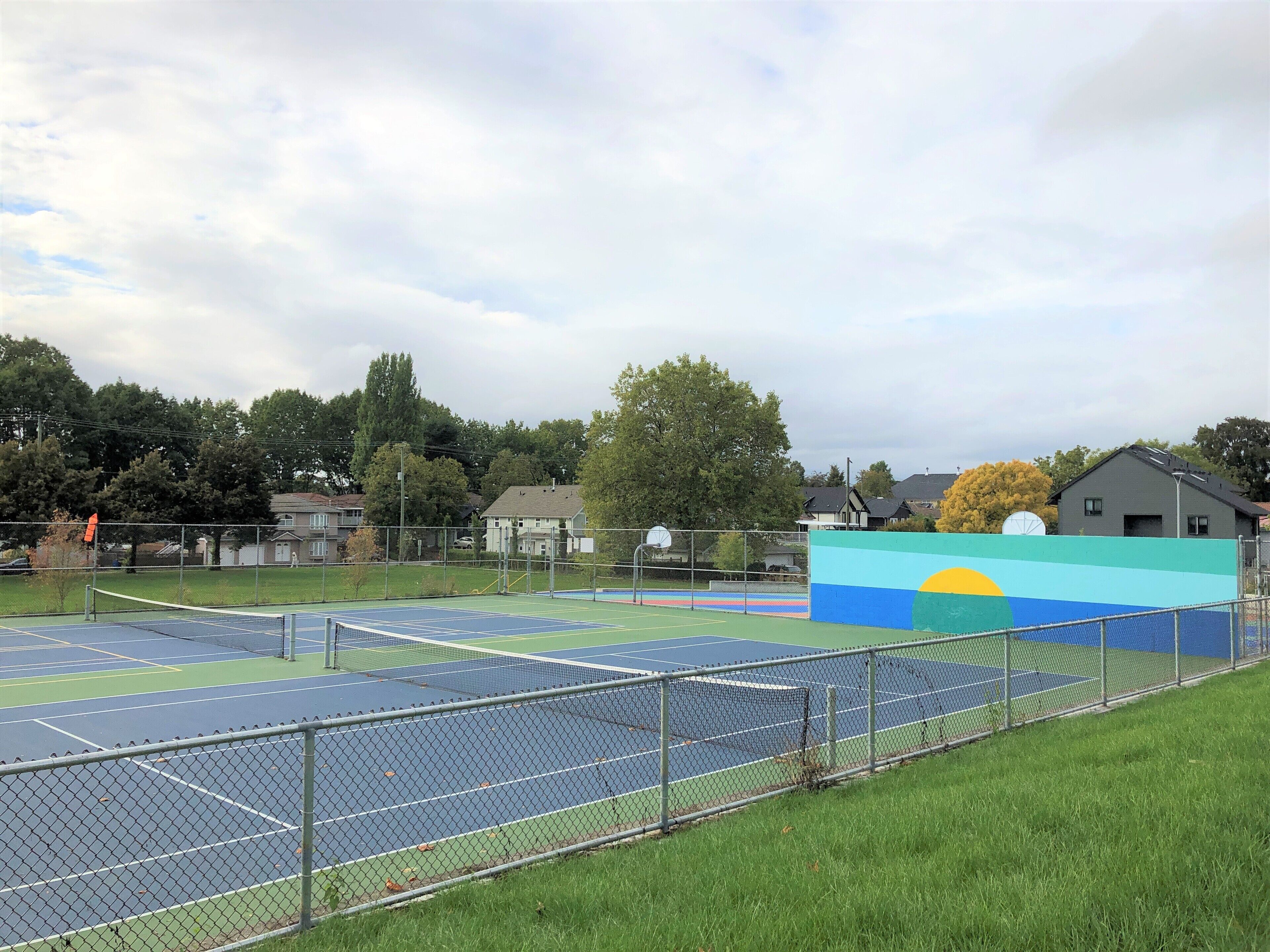 An outdoor tennis court with a fence, a basketball court, and a mural.