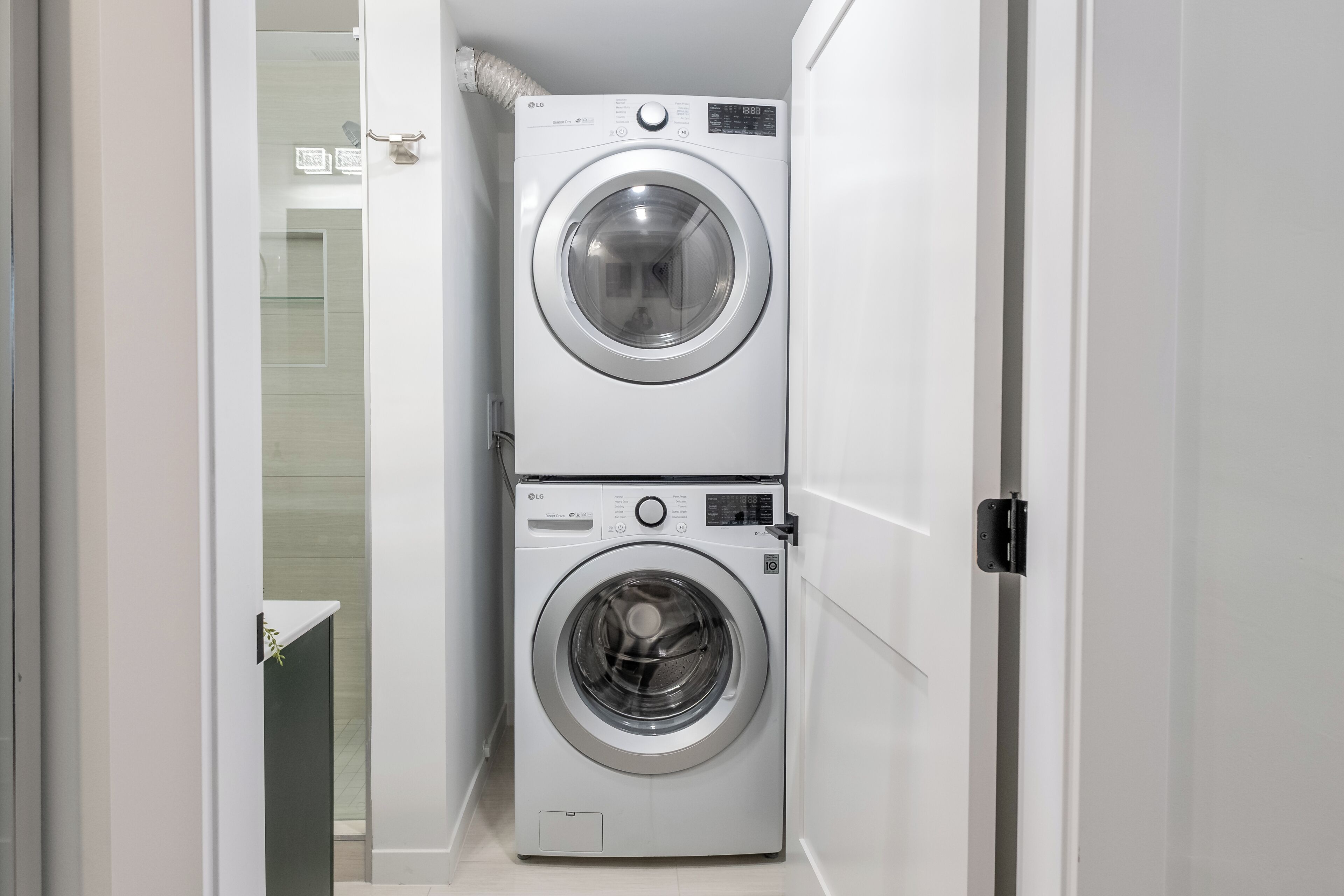 A compact laundry room with two stacked washing machines, a glass door leading to a bathroom, and a white tiled floor.