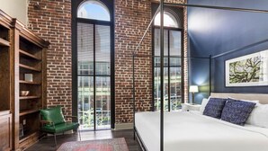 Un dormitorio con pared de ladrillo, un ventanal grande, una silla verde, una cama con ropa de cama blanca y una alfombra estampada.