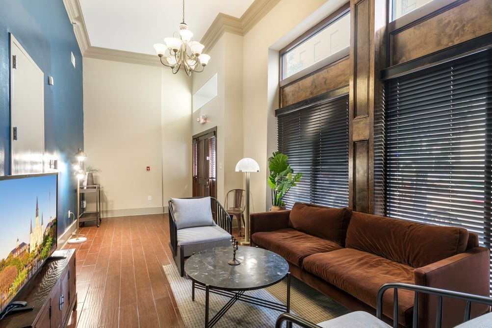 A living room with a brown sofa, a round coffee table, and a TV on the left. There is a chandelier hanging from the ceiling and a window with blinds on the right.