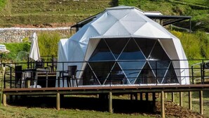 Estructura de domo geodésico con un porche cubierto, mobiliario exterior y vistas a una colina.
