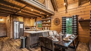 L’intérieur d’un chalet rustique en bois comprend une cuisine et un coin repas, avec un grand îlot central, un plancher en bois et une vue sur l’extérieur par une fenêtre.