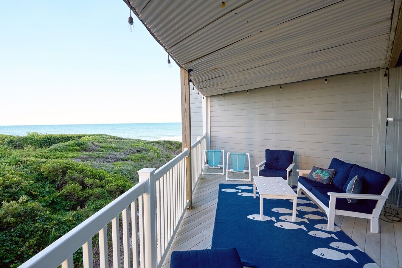 Varanda coberta com vista para o oceano, com área de estar com duas cadeiras e uma mesa, e um tapete azul com motivos de peixes.