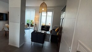 Una sala moderna con un sofá, una mesita de centro y un lámpara colgante. Hay un televisor montado en la pared y una ventana con persianas.