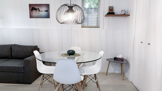 A modern living room with a glass table, white chairs, a gray sofa, a pendant light, and a window with blinds.
