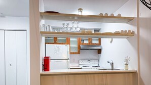 A compact kitchen with a refrigerator, stove, and sink. There are wooden shelves above the counter holding dishes and glasses.