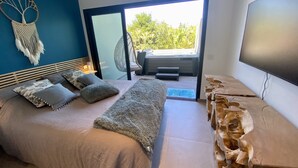 Un dormitorio moderno con una cama grande, vista a zonas verdes a través de una puerta corrediza, un televisor montado en la pared y una escultura de madera original.