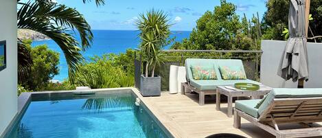 A poolside area with a view of the ocean, lounge chairs, and a table with a glass.