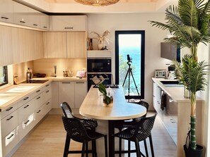 Une cuisine moderne avec un îlot central, des chaises noires et une vue sur la mer grâce à une grande fenêtre.