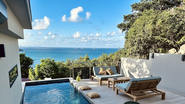 Zona de piscina con sillones de descanso y una mesa, con vista a un paisaje marino.