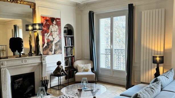 A living room with a fireplace, a sofa, a coffee table, a bookshelf, and a large window with curtains.