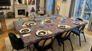 A dining area with a table set for six, surrounded by chairs, in a room with a fireplace, bookshelves, and a large window.