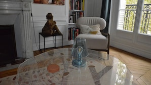A room with a fireplace, a bookshelf, a white armchair, and a round glass table with a decorative glass object.