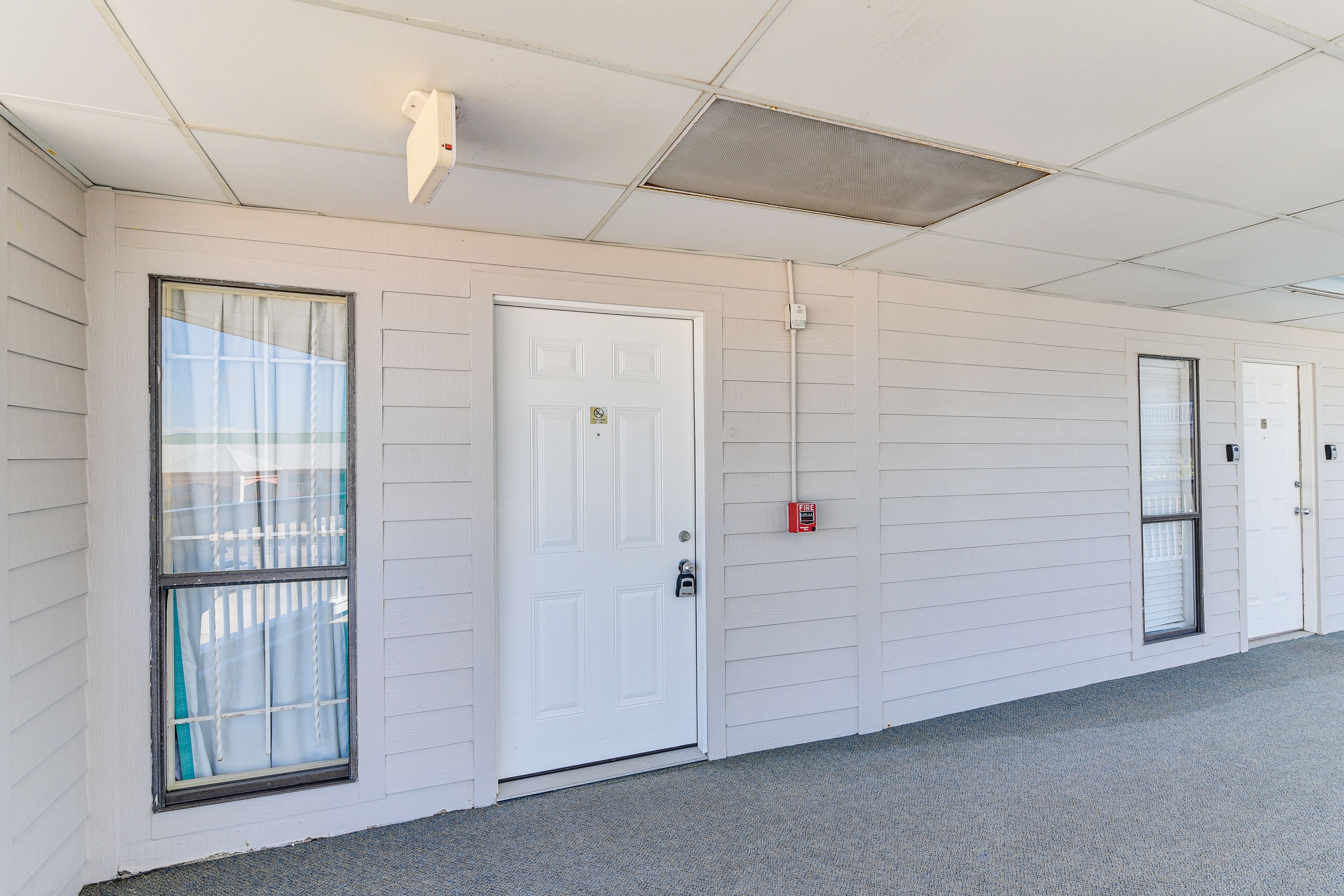 A corridor with white doors, a window with curtains, and a fire alarm on the wall.