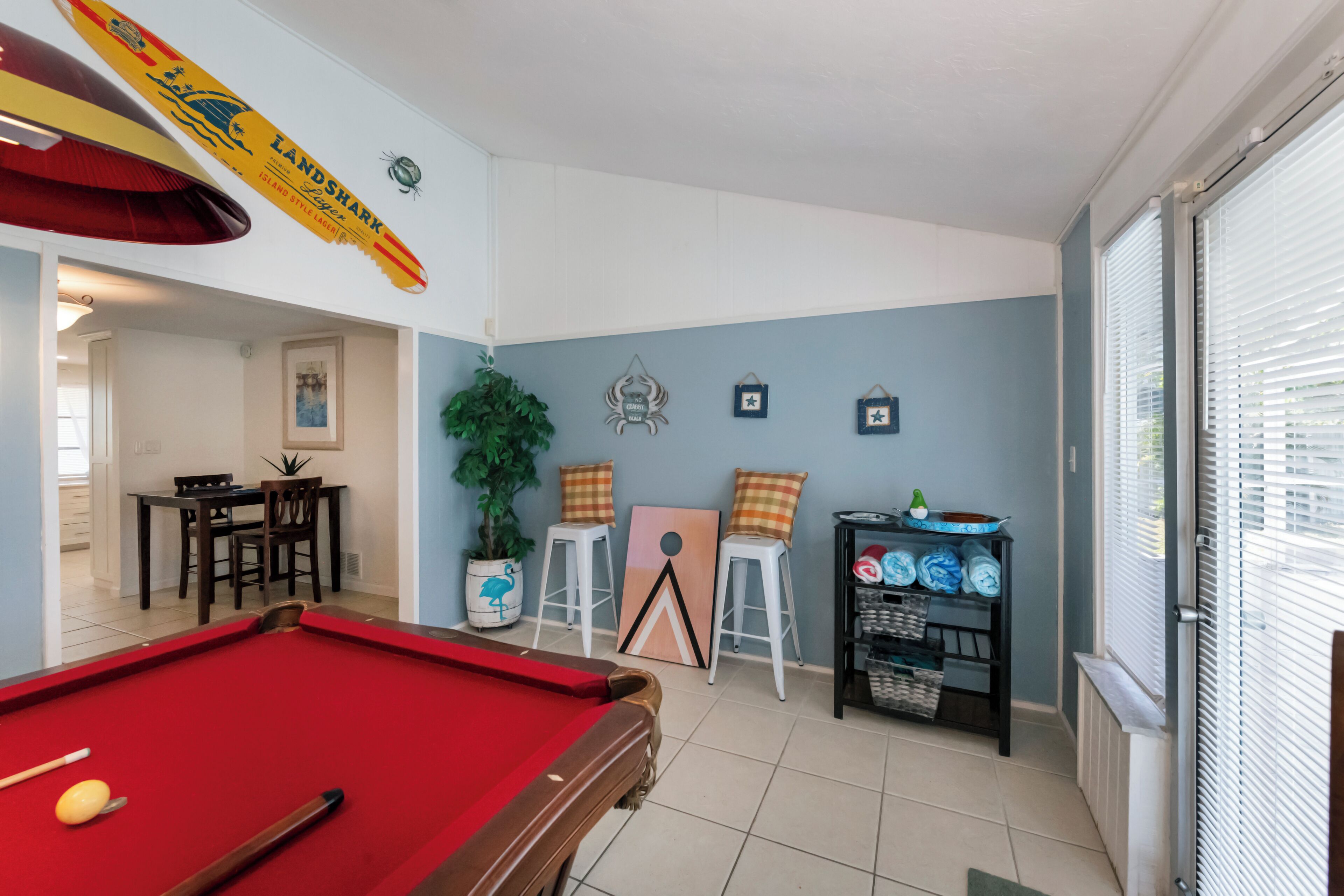 A pool table in a room with a dining area, a plant, and a shelf with bowls.
