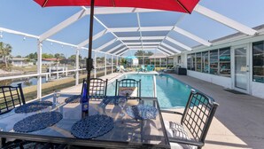 A covered outdoor pool area with a glass table, chairs, and a red umbrella. - Heated Pool, Canal Front, Kayaks - Paradise Awaits!  (Port Charlotte)
