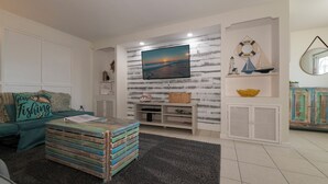 A living room with a TV mounted on a textured wall, a sofa with a 'Gone Fishing' pillow, a coffee table with books, and a small shelf with decorative items. - Heated Pool, Canal Front, Kayaks - Paradise Awaits!  (Port Charlotte)
