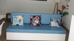 Um sofá azul com almofadas florais, uma luminária e uma planta em vaso num quarto com janela e obras de arte na parede.
