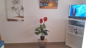 Uma sala de estar com piso de azulejos, um vaso com flores vermelhas, um quadro de palmeiras, um quadro com uma cena de praia e uma televisão exibindo um mapa.