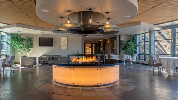 A modern lounge area with a central fire feature, seating arrangements, and large windows providing natural light.
