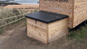 A wooden building labeled "WILD SCOTTISH SAUNA" with a black roof, situated on a beach with the ocean in the background. - Captivating 2-bed Cottage in Kingsbarns (St. Andrews)