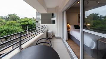 A modern balcony with a table and chairs, a bed with white bedding, and a view of greenery and buildings.