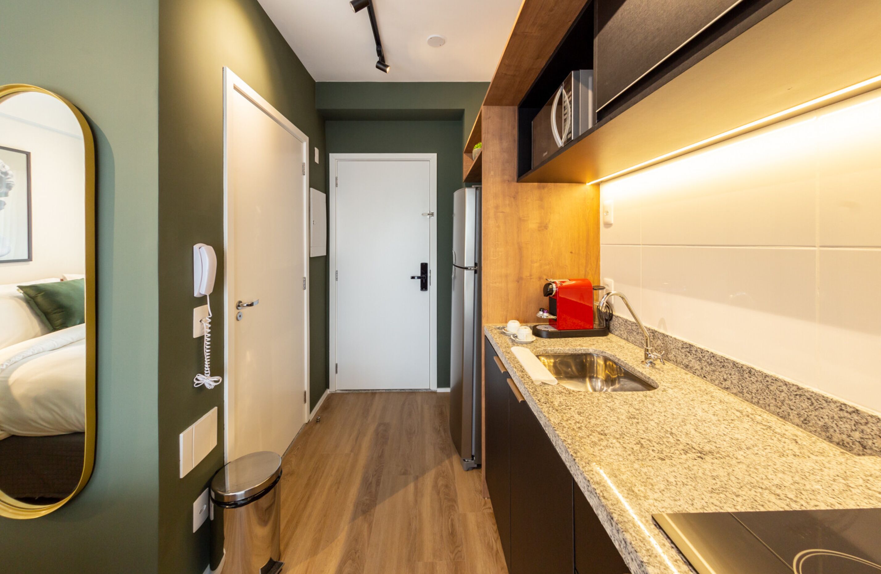 Un pasillo estrecho con piso de madera, una puerta blanca y una zona de cocina con encimera de granito y pileta.