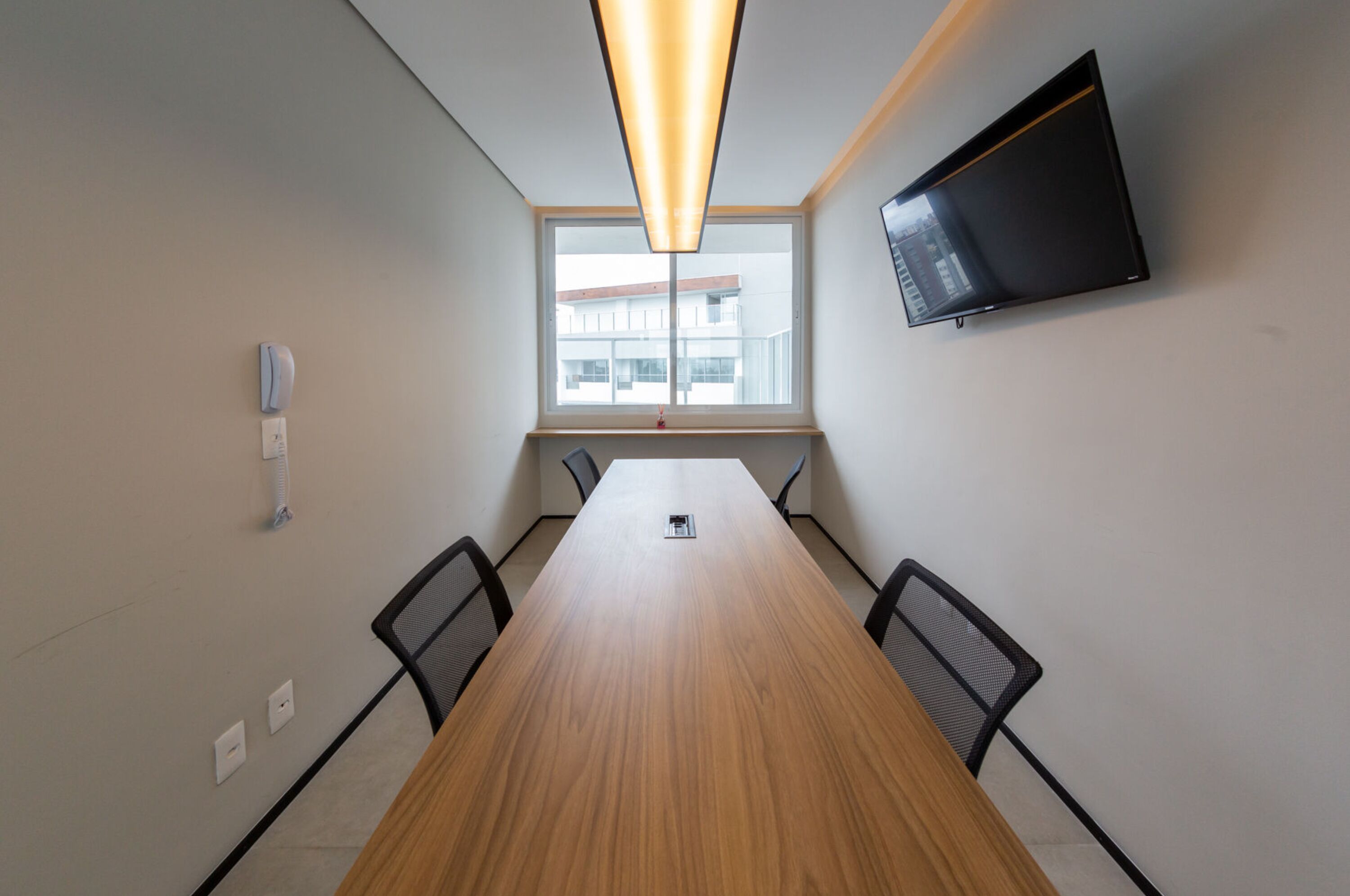 A conference room with a long wooden table, four chairs, a mounted TV, and a window with a view of buildings.