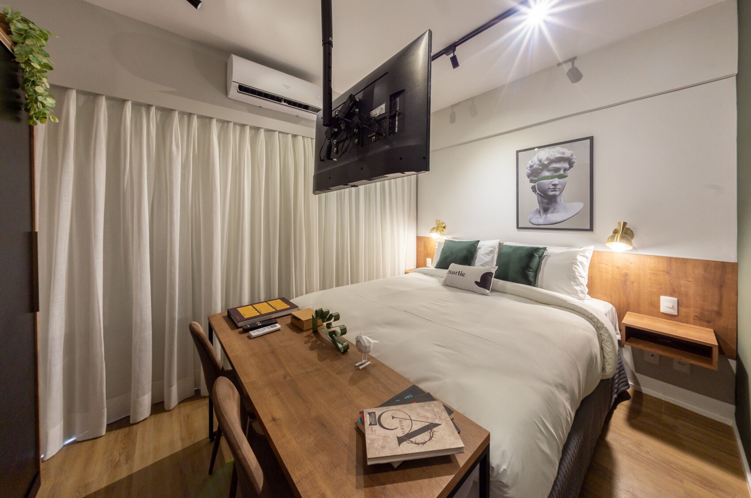 A modern hotel room with a neatly made bed, a wooden desk with a chair, a flat-screen TV mounted on the wall, and a framed artwork above the bed.