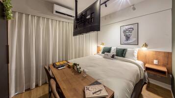 A modern hotel room with a neatly made bed, a wooden desk with a chair, a flat-screen TV mounted on the wall, and a framed artwork above the bed.