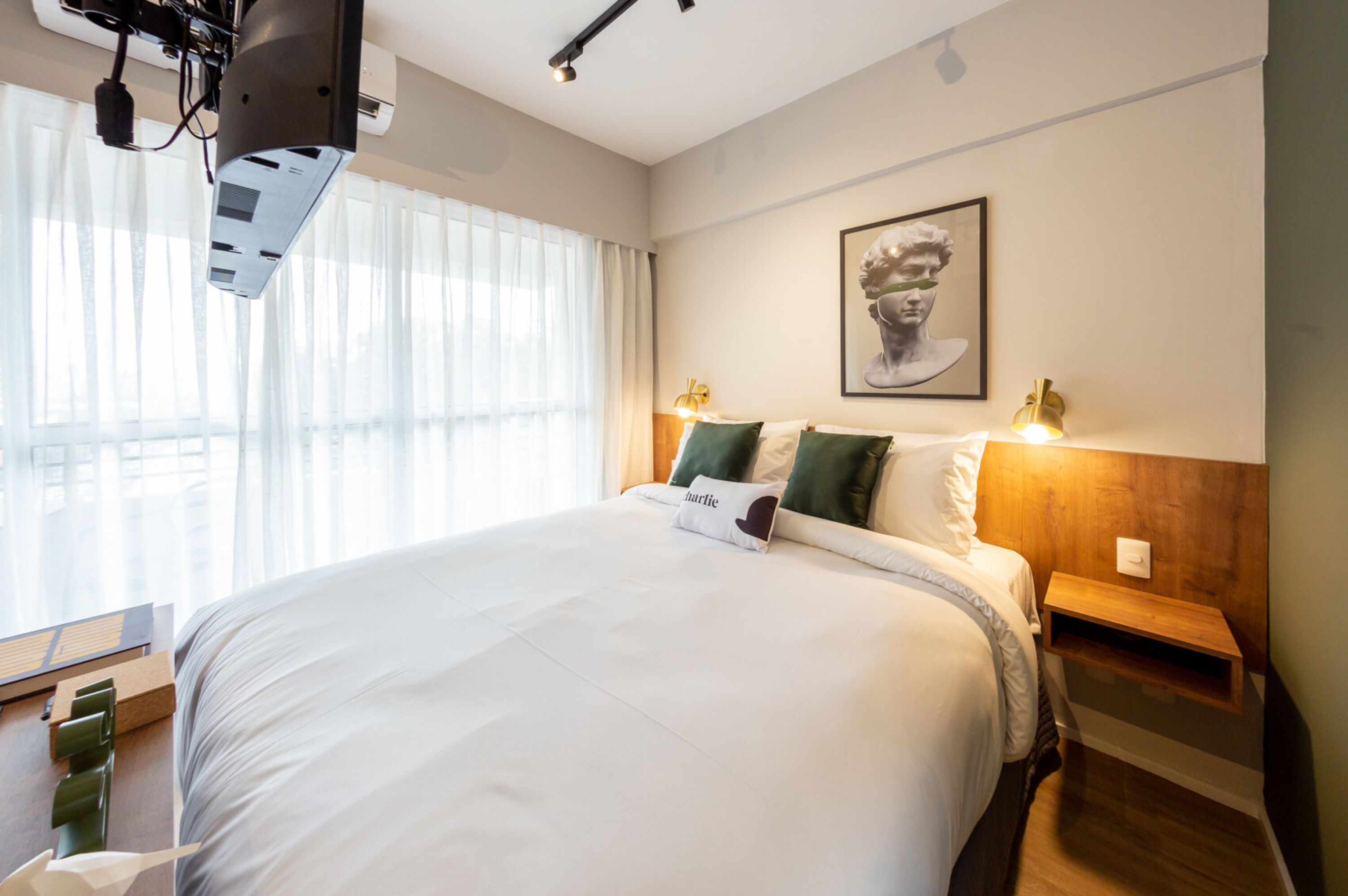 Una habitación de hotel con una cama grande, una mesita de noche de madera, un estante en la pared y una obra de arte enmarcada sobre la cama.