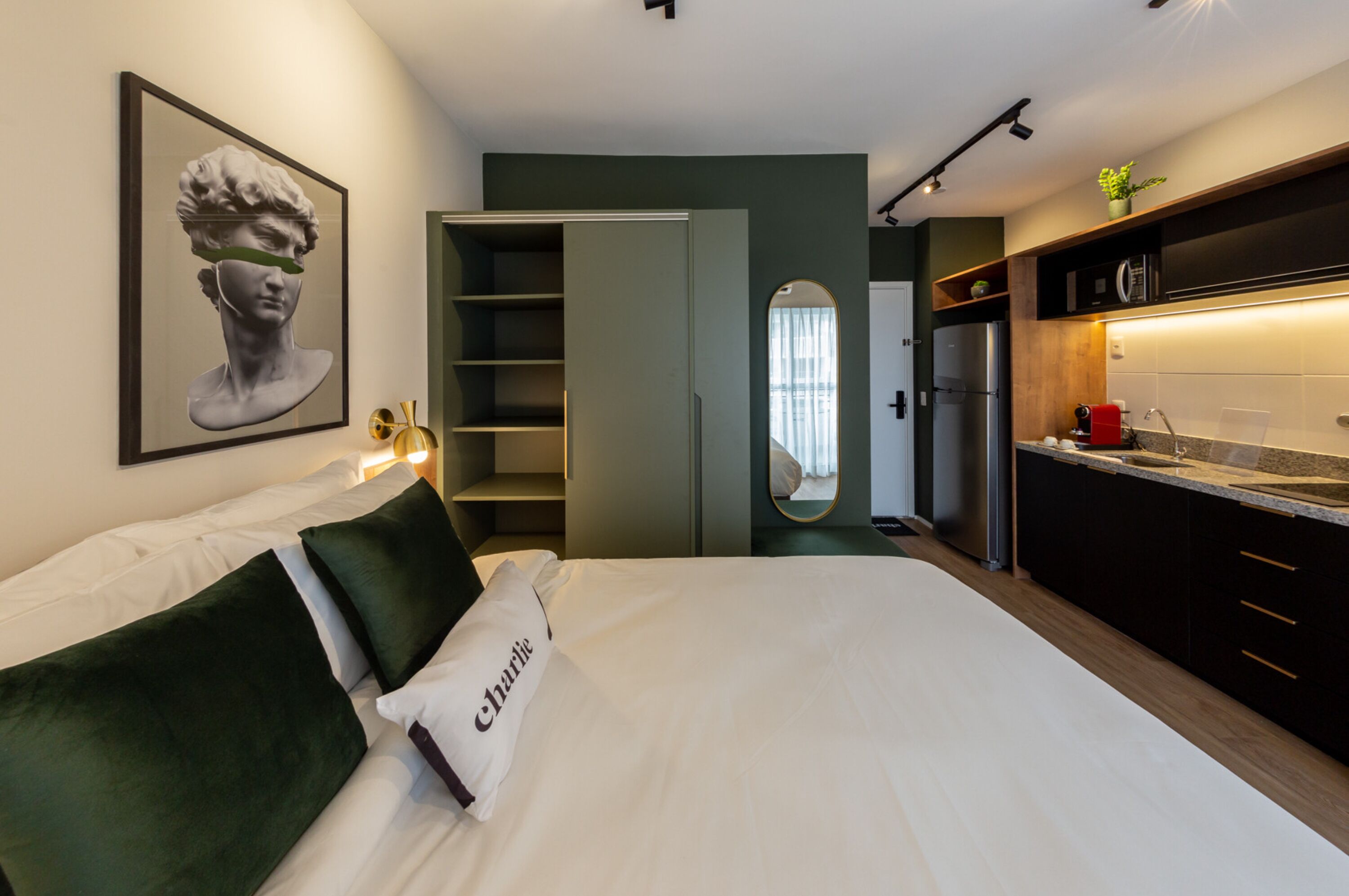 Et moderne hotelværelse med seng, grøn pude, indrammet kunstværk, et skab, et spejl og et tekøkken med køleskab og vask.