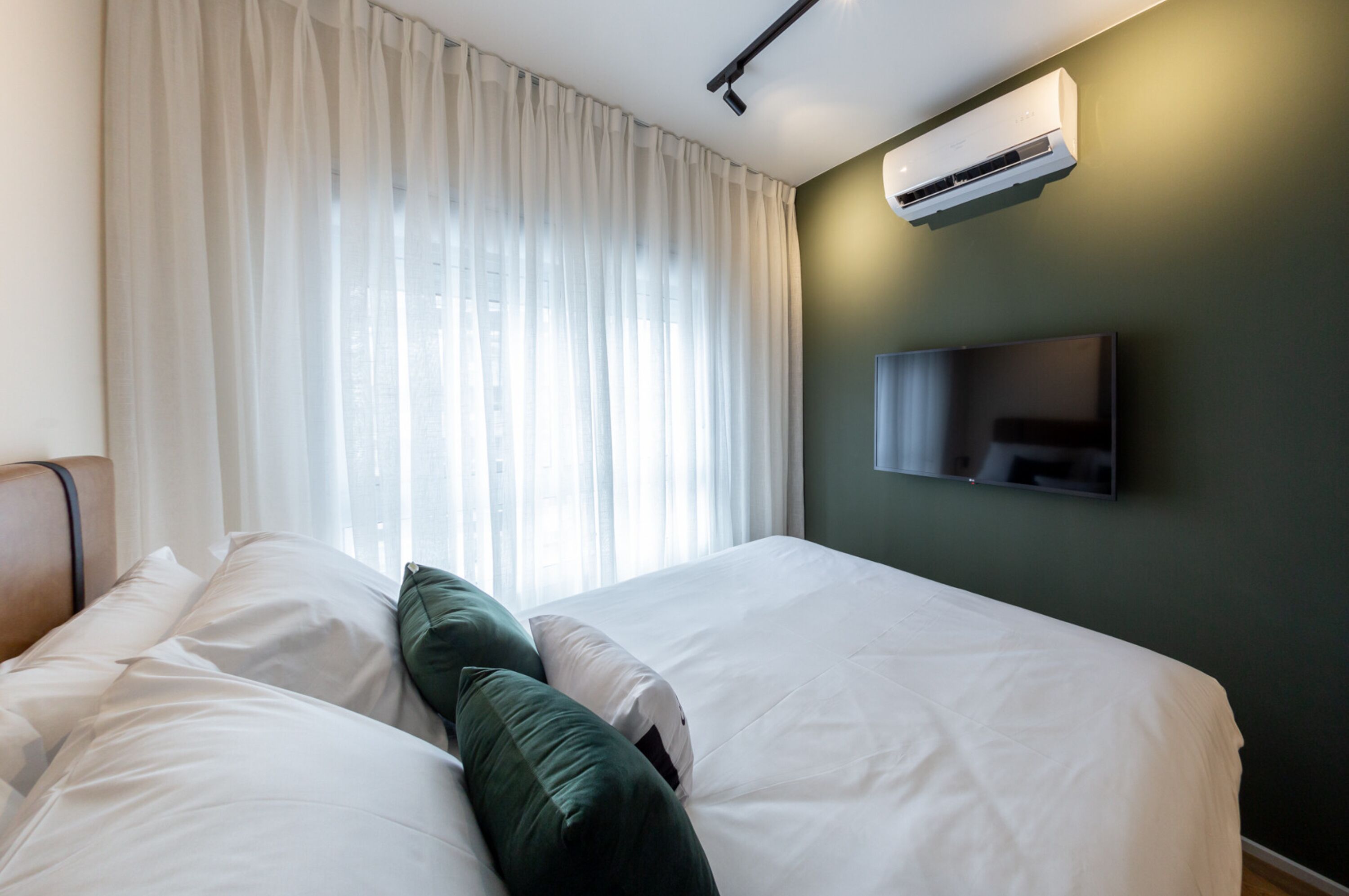 A hotel room with a bed, white curtains, a mounted television, and an air conditioning unit.