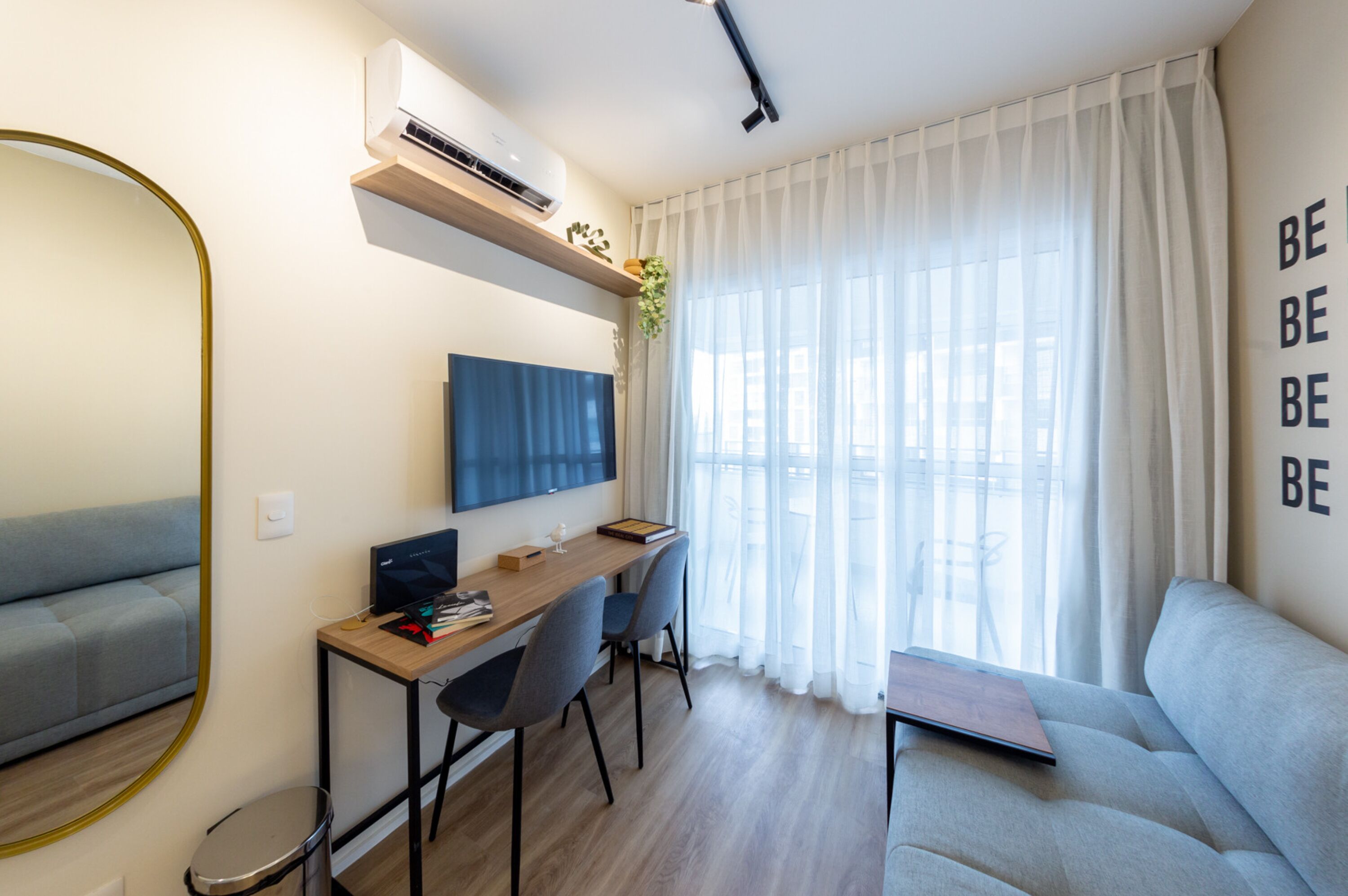 Una sala de estar moderna con un sofá, un escritorio con una computadora, un televisor y un amplio ventanal con cortinas translúcidas.