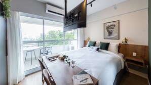 Una habitación de hotel moderna con una cama grande, un escritorio con silla, un televisor montado y un balcón con mesa y sillas.