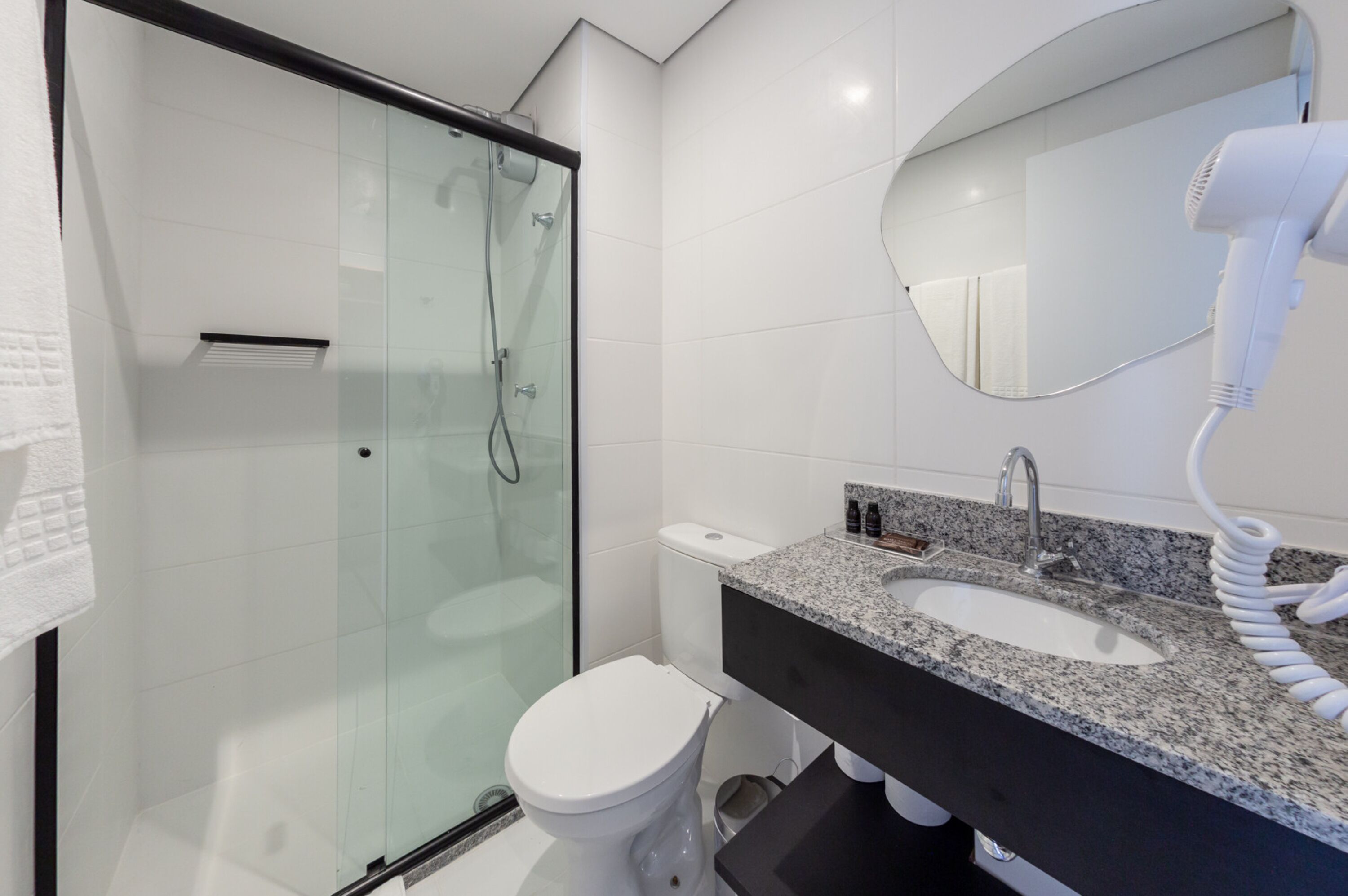 Un baño moderno con ducha envuelta en vidrio, inodoro blanco, lavabo con encimera de granito y secador de pelo.