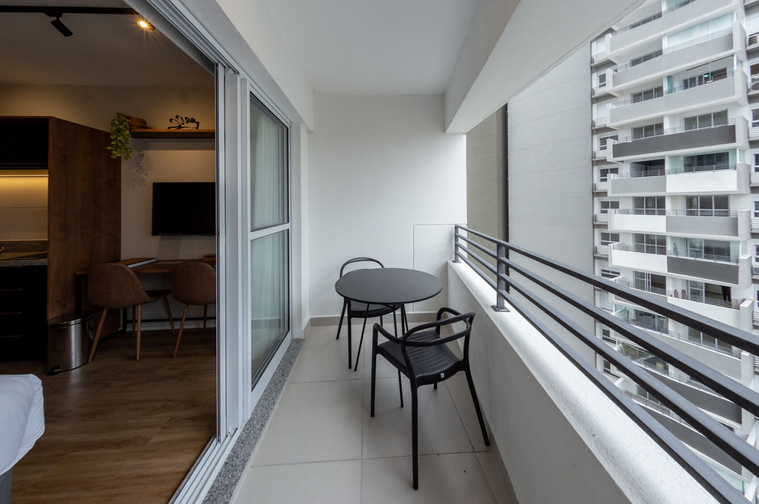 A modern apartment with a balcony, a dining area with two chairs, a kitchen with a TV, and a large window overlooking a building.