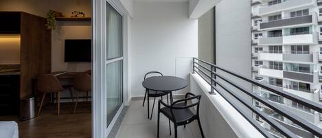 A modern apartment with a balcony, a dining area with two chairs, a kitchen with a TV, and a large window overlooking a building.