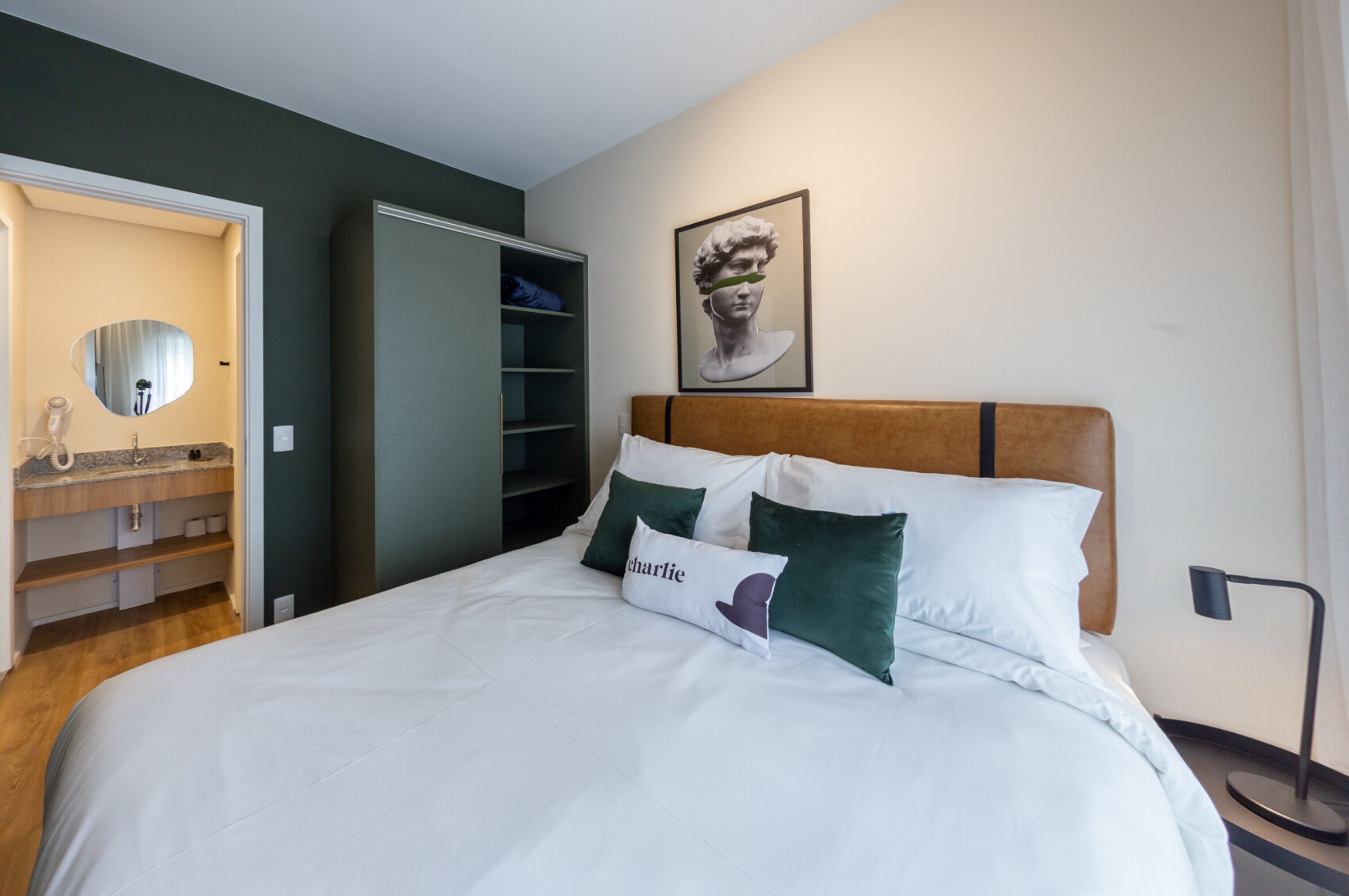 Una cama bien hecha con ropa de cama blanca y verde, un cabecero de madera y un almohadón con el nombre 'Charlie'.