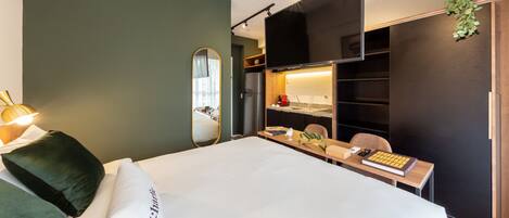 A modern hotel room with a large bed, a desk with a computer, a chair, and a wardrobe.