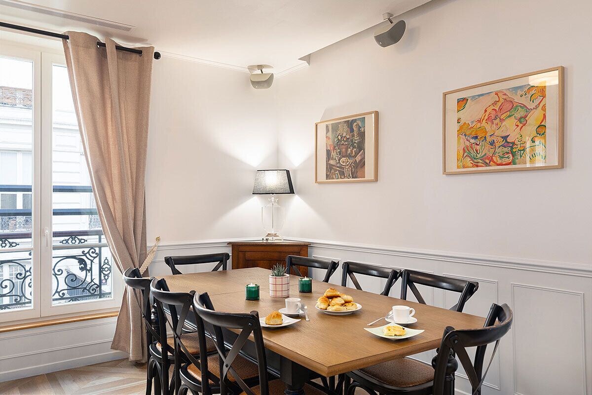 Een eetgedeelte met een houten tafel, zwarte stoelen en een raam met gordijnen. Twee ingelijste kunstwerken hangen aan de muur en er staat een lamp op de tafel.