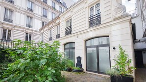 Une cour avec un grand arbre, une fontaine décorative et un bâtiment doté d’une porte en verre et de volets noirs.