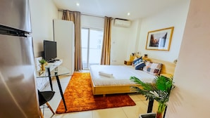 Quarto de hotel com cama, escrivaninha com computador, televisão, janela com cortinas e uma planta.