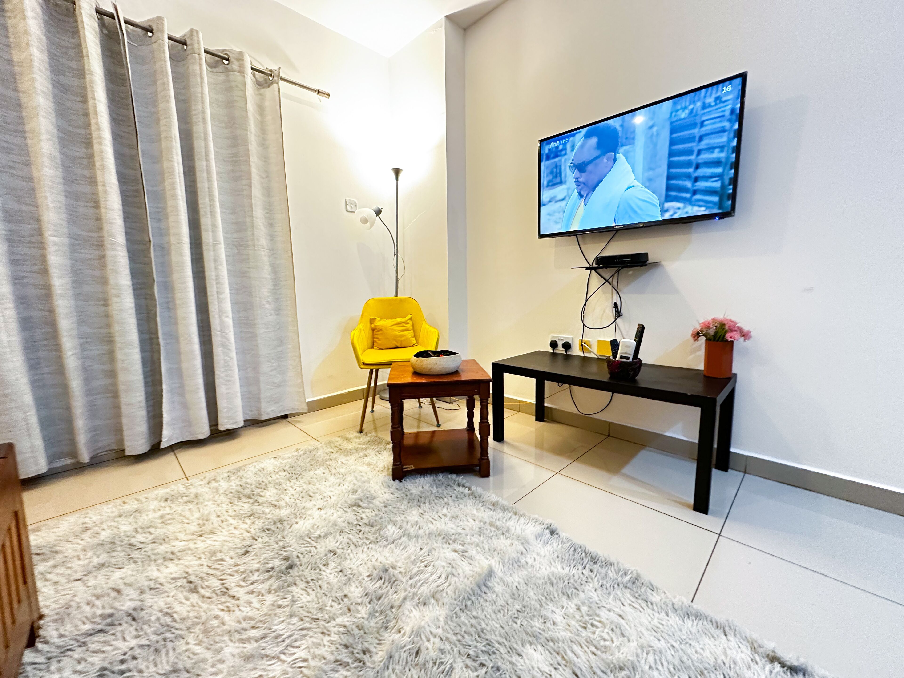 Un salon avec un téléviseur fixé au mur, un fauteuil jaune, une petite table et un tapis moelleux.