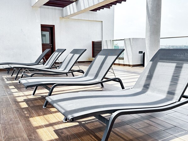 A rooftop terrace with multiple lounge chairs arranged in a row, a wooden floor, and a view of the cityscape.
