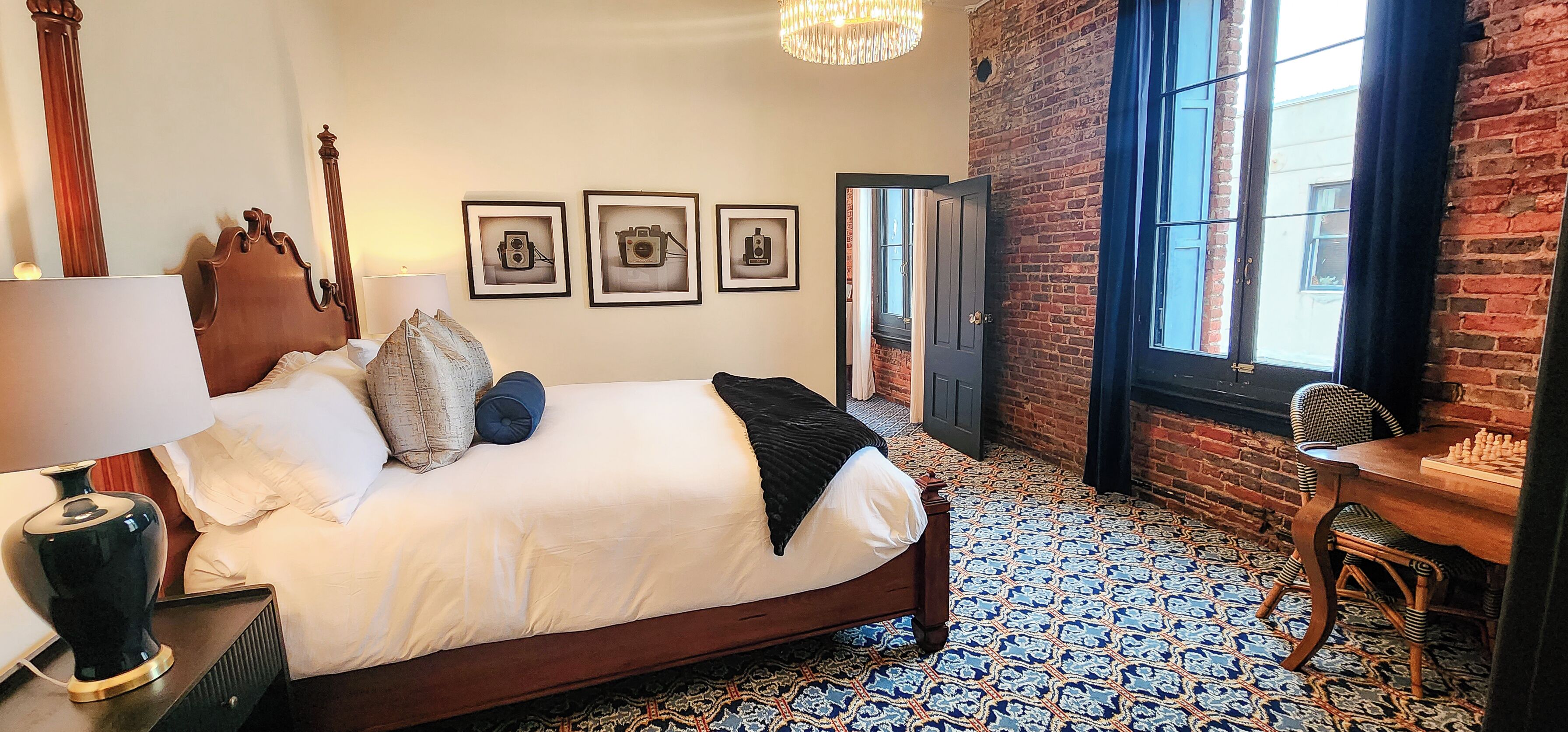 Quarto de hotel com uma cama, uma escrivaninha com peças de xadrez, uma luminária, uma cadeira e piso estampado.
