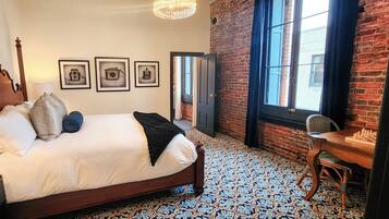 Quarto de hotel com uma cama, uma escrivaninha com peças de xadrez, uma luminária, uma cadeira e piso estampado.