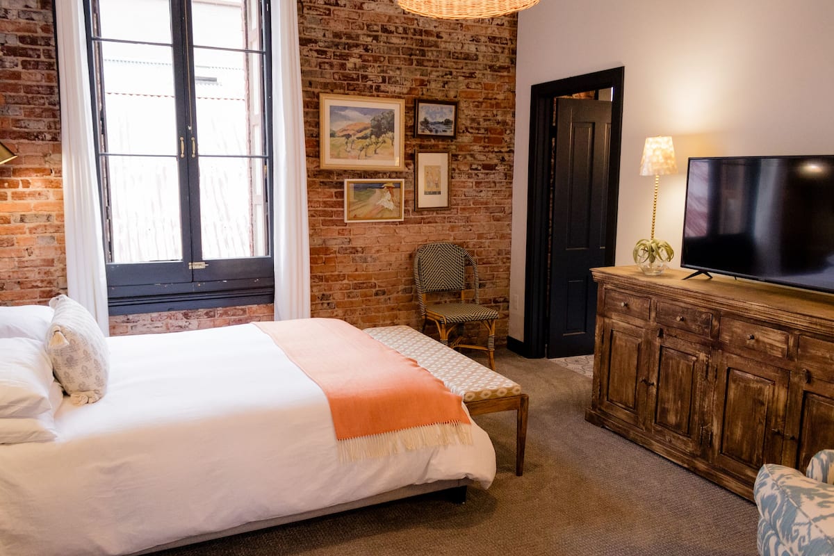 Quarto com uma cama grande, um banco de madeira, uma televisão sobre um armário de madeira e uma parede de tijolos com quadros.